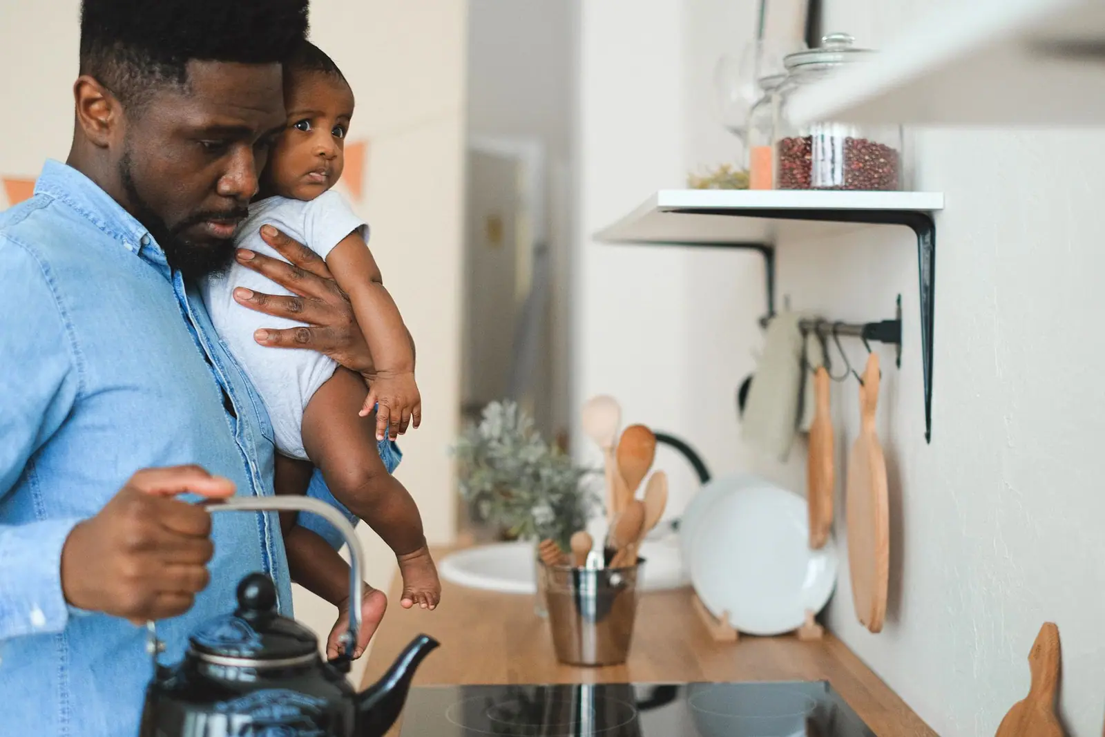 An adult holding a baby beside a kitchen counter and kettle