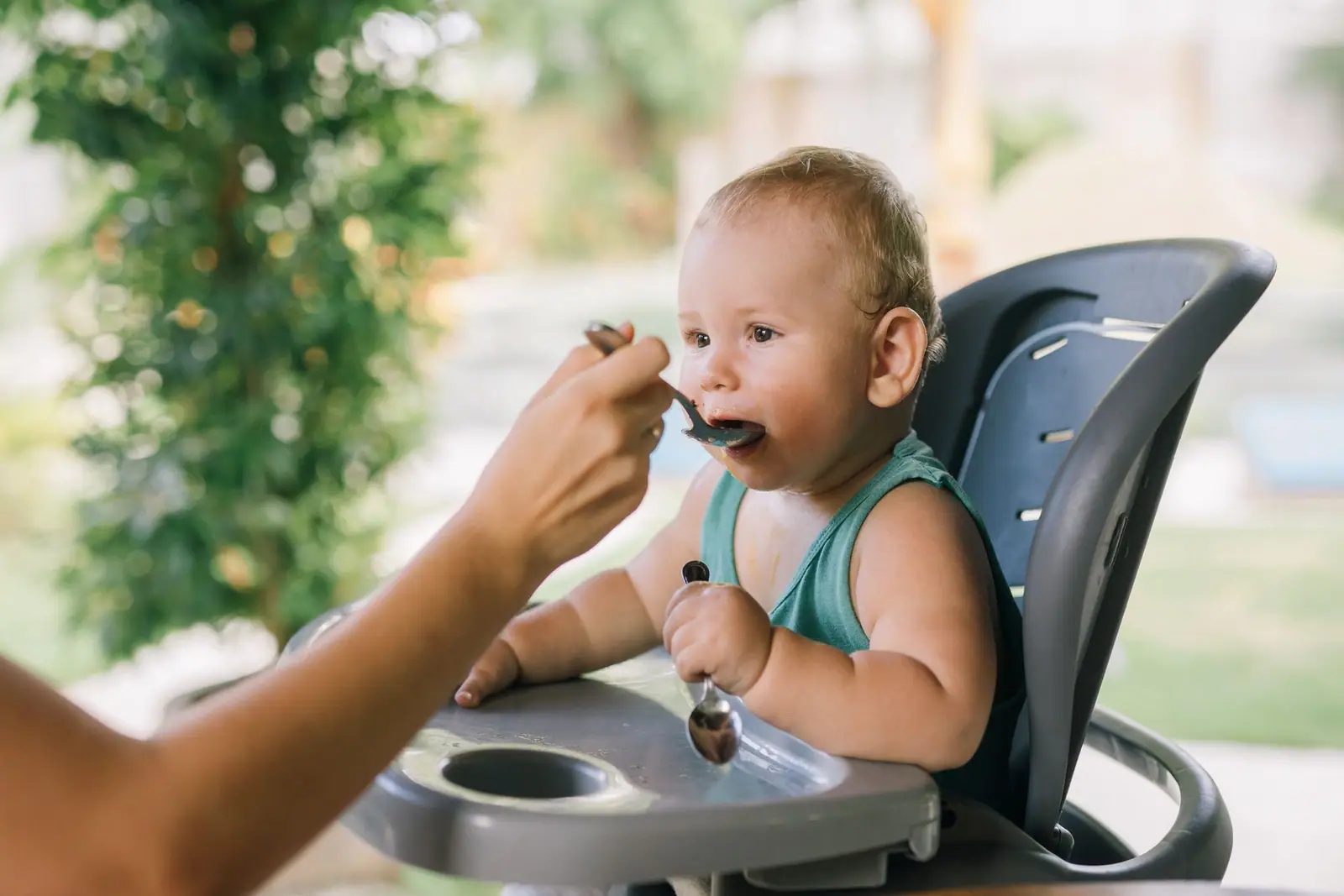 An adult spoon-feeding a child seated in a black high chair outdoors